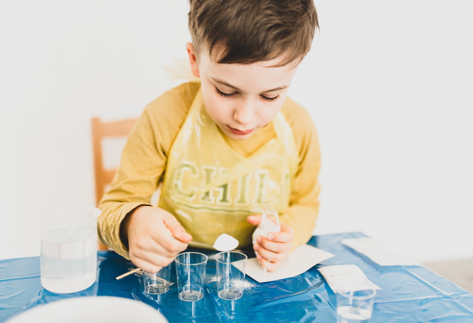 A young boy participates in an age-appropriate STEM activity with small cups and measuring spoons. He has covered himself with an apron and his table with a covering for safety purposes.