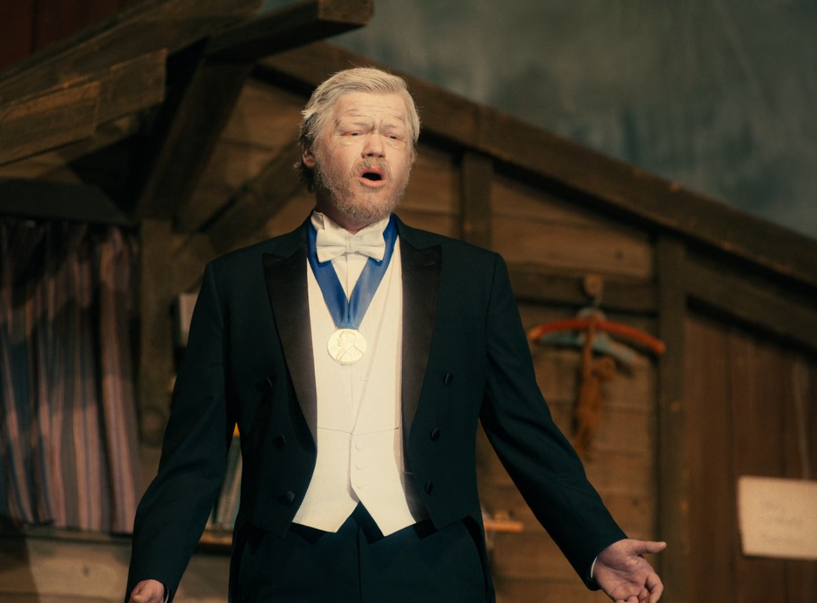 Jesse Plemons in I'm Thinking of Ending Things (2020). An elderly Jake in ageing makeup with grey hair, is wearing a tuxedo and a Nobel Prize medal, performing a song on stage, gazing out at the audience.