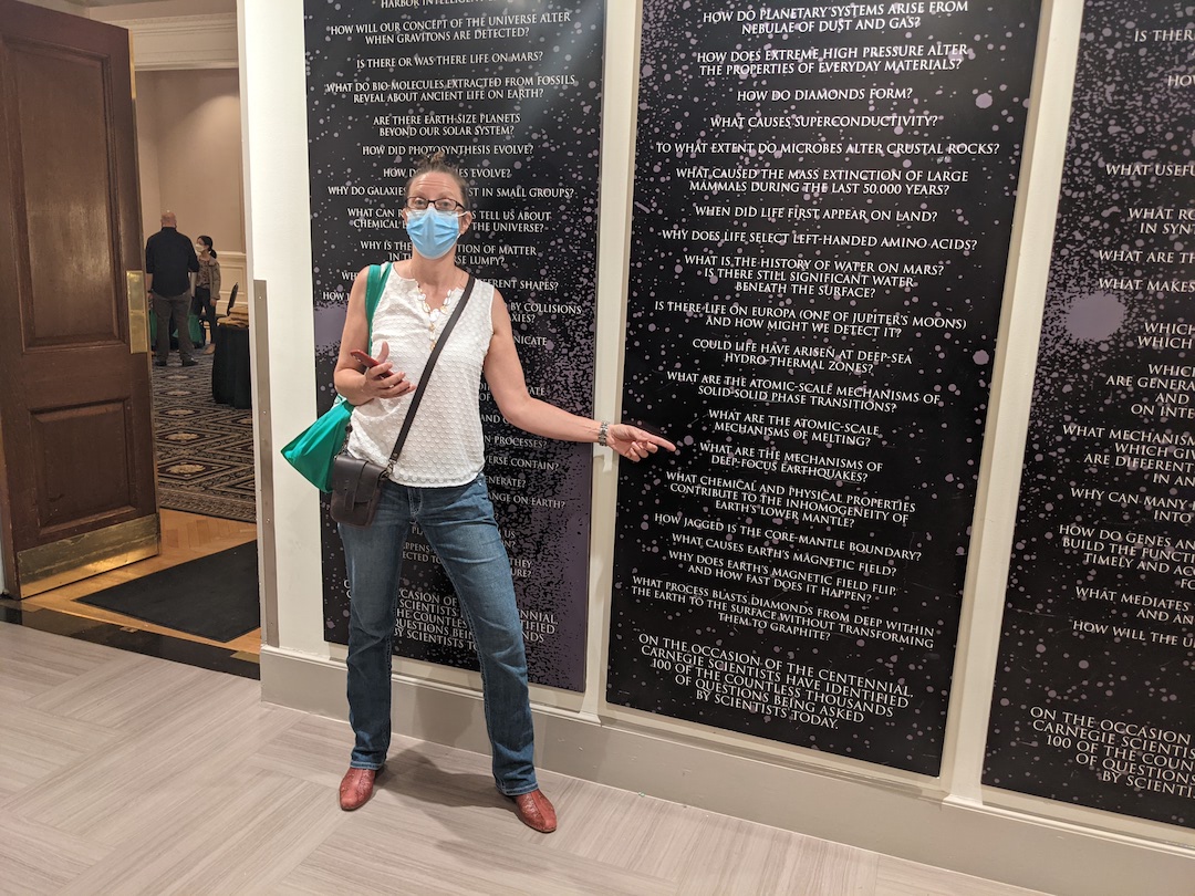 Lara Wagner stands next to a set of questions hanging at the P-Street office including "What is the source of deep focus earthquakes?"
