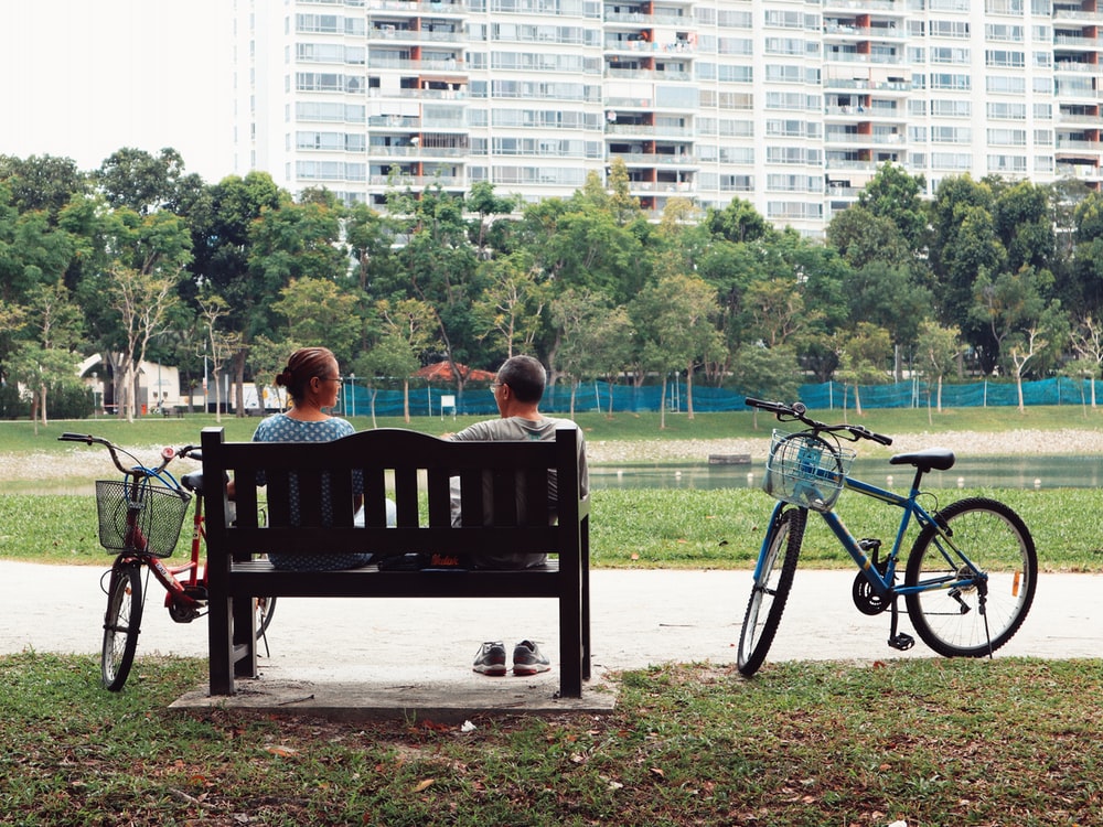 man and woman sitting on bench near two bikes viewing green field during daytime