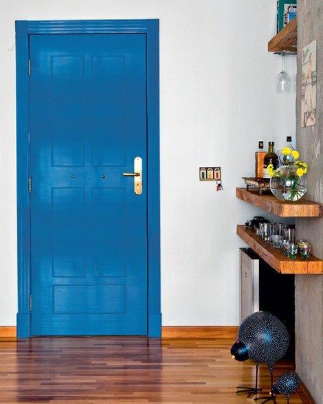 Porta de entrada com porta azul royal, piso de madeira, parede com prateleiras de madeira com bebidas, taças e frigobar.
