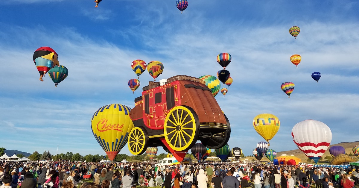 People crowd an open field at a mass hot air balloon launch, with a big Wells Fargo wagon hot air balloon in the middle.