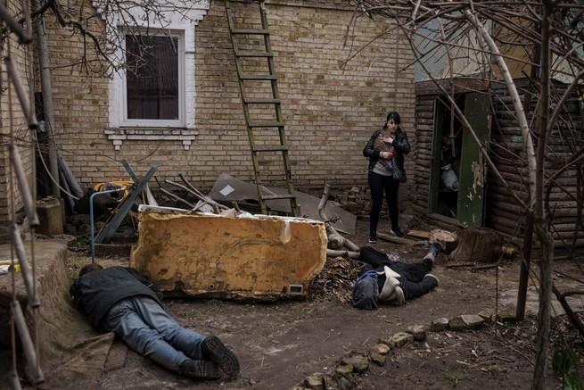 color photo of a woman holding a cat in a ruined yard by a house and looking at two corpses lying face down on the ground
