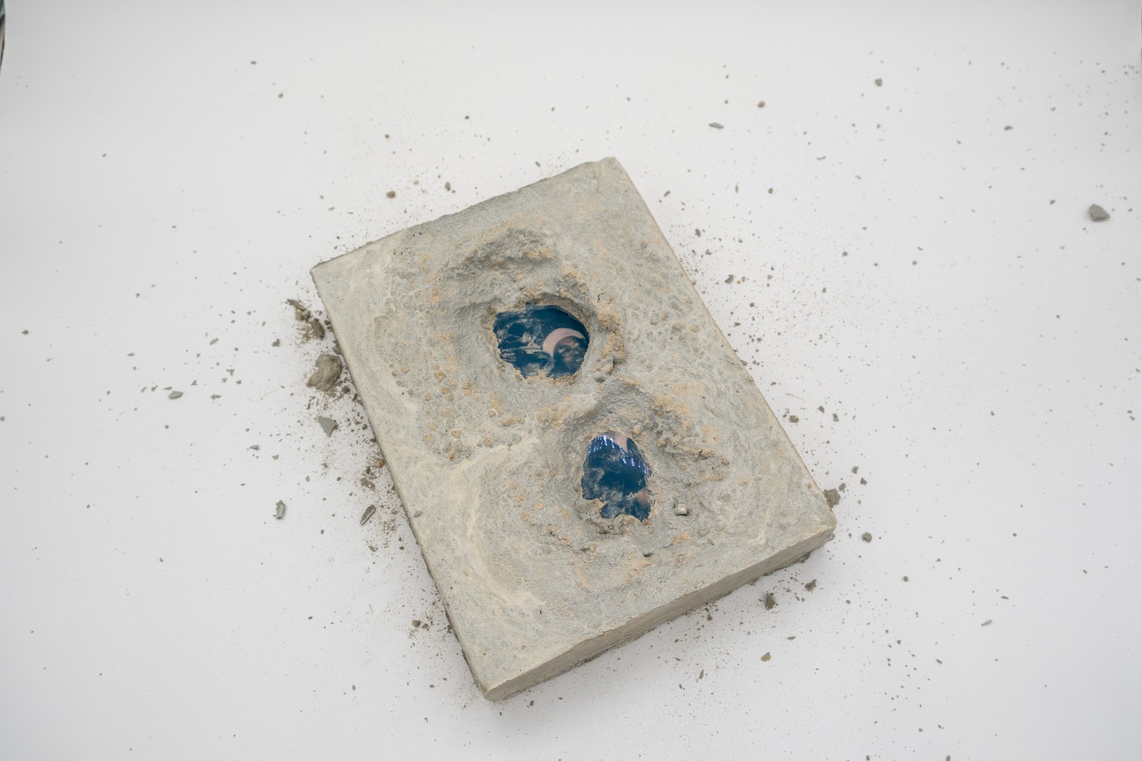 Image: A view of the 2002 work, Untitled no. 11. The work is a rectangle of concrete with small fragments and residue crumbling from it. A blue-hued photo of a person is encased by the concrete but there are two holes dug into the concrete, revealing details of the photograph. Photo courtesy of Sibyl Gallery.
