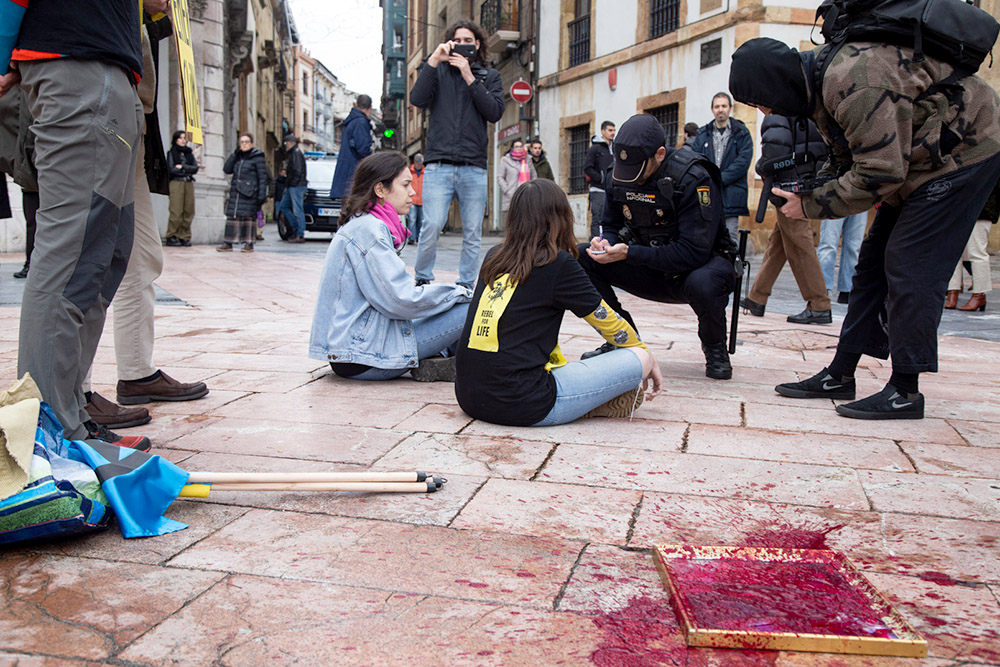 Two rebels sit on pavement, the painting covered in red paint beside them, while a policeman asks them questions