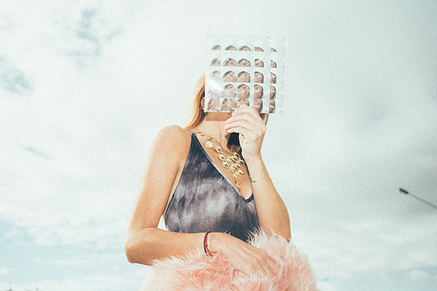 Model standing against a sky background wearing a gray tank top. she holds a piece of glass in front of her face, which produces multiple views of her face