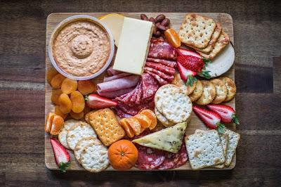 A charcuterie board with fruit, meats, crackers, chips, dips, seeds, nuts, cheeses