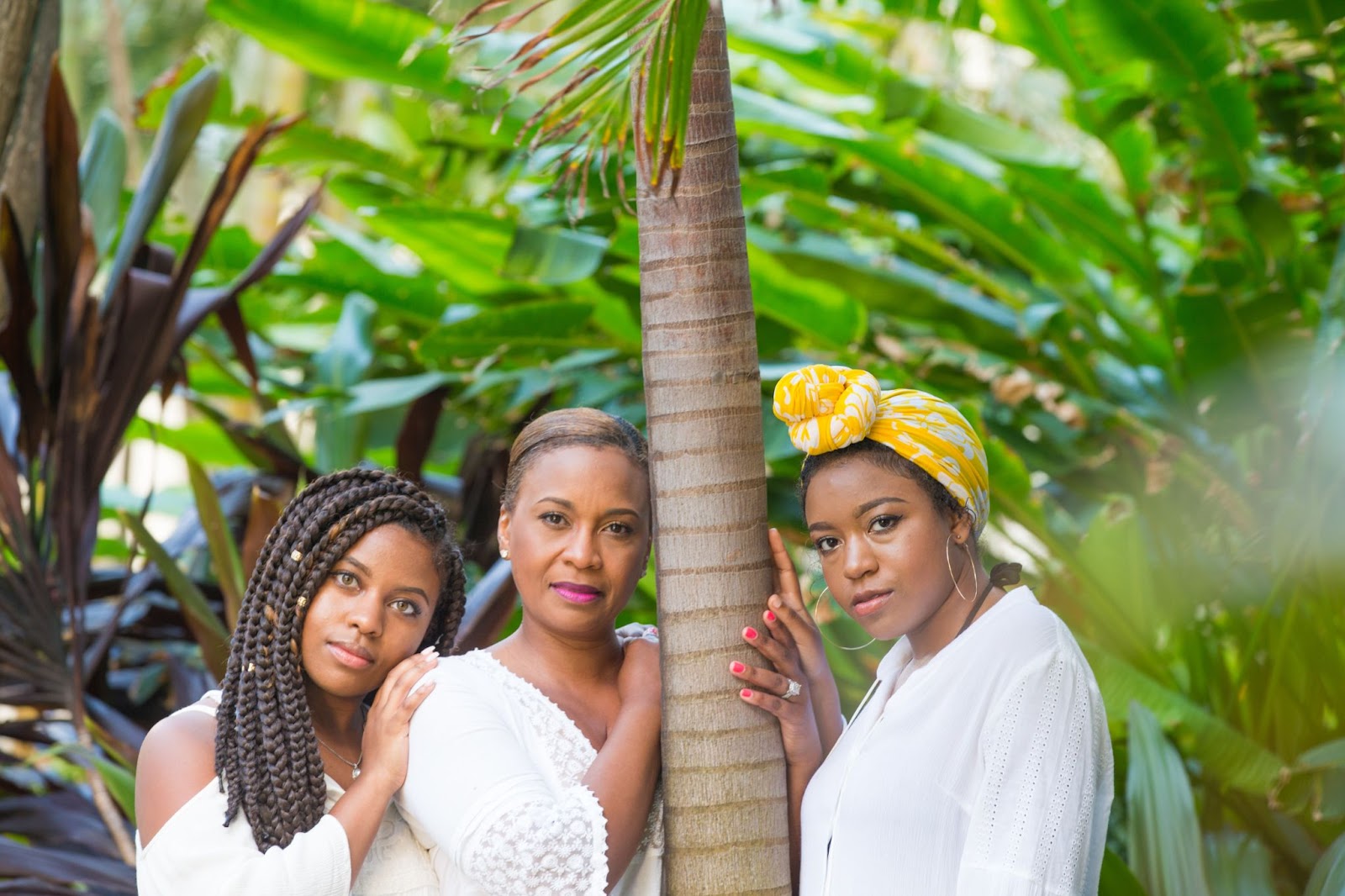 Three Black women pose around a palm tree in a tropical rainforest.