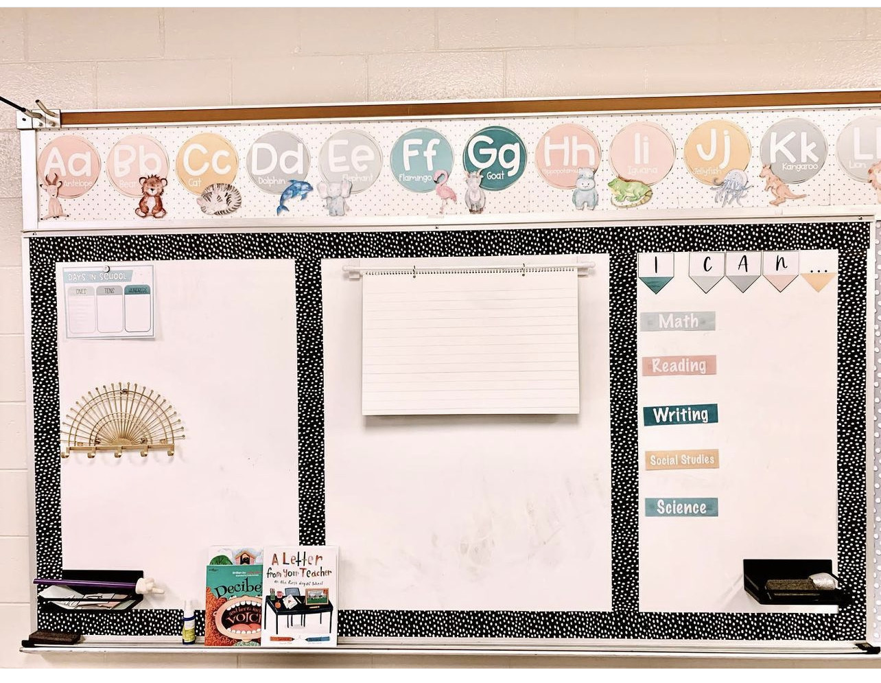 This image shows a whiteboard that has been cut into three sections using a black with white polka dot border. The section on the far left holds math materials. The middle section holds a large lined pad of paper. The right section holds I Can statements for five different subjects. 