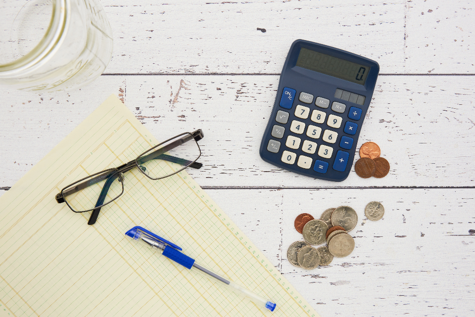 Investment ready: uma folha de papel, calculadora, óculos, caneta e algumas moedas sobre uma mesa de madeira branca