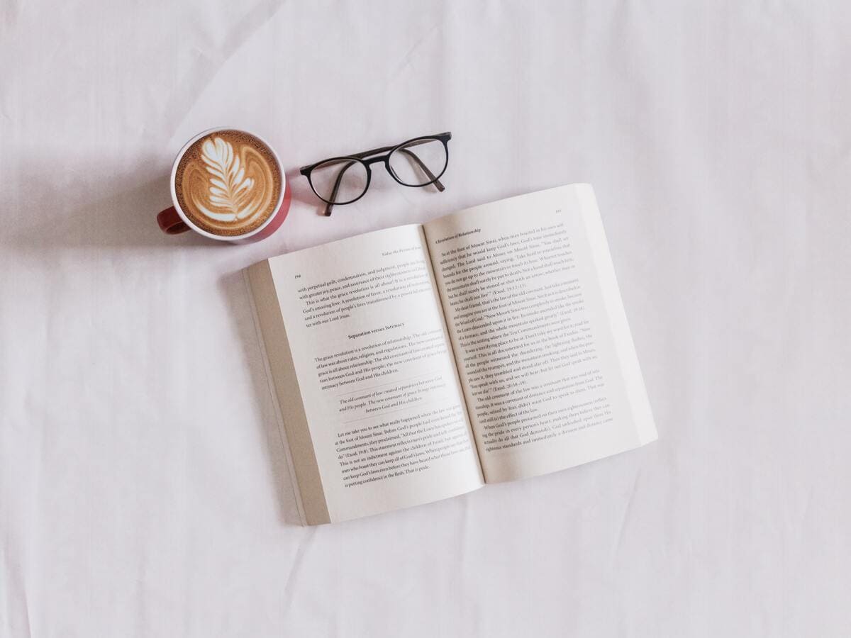 Un libro aperto, degli occhiali e un cappuccino posti su una superficie bianca