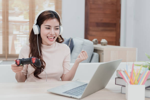 A beautiful Asian woman is carrying a joystick to play games. Go online via the internet using your home laptop as entertainment. A new normal life A beautiful Asian woman is carrying a joystick to play games. Go online via the internet using your home laptop as entertainment. A new normal life gaming laptop stock pictures, royalty-free photos & images