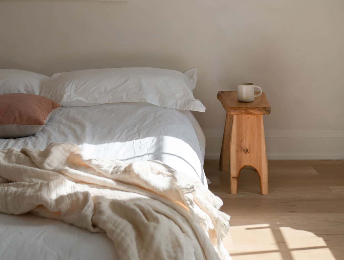A bed with no headboard covered in linen bedding with a wooden stool beside.