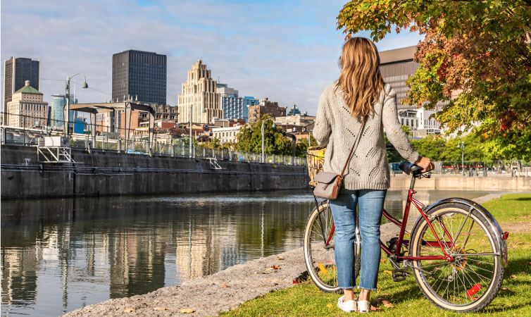 A woman is standing beside her bike on a sunny day, looking across the water at the city skyline of Montreal, QC. 