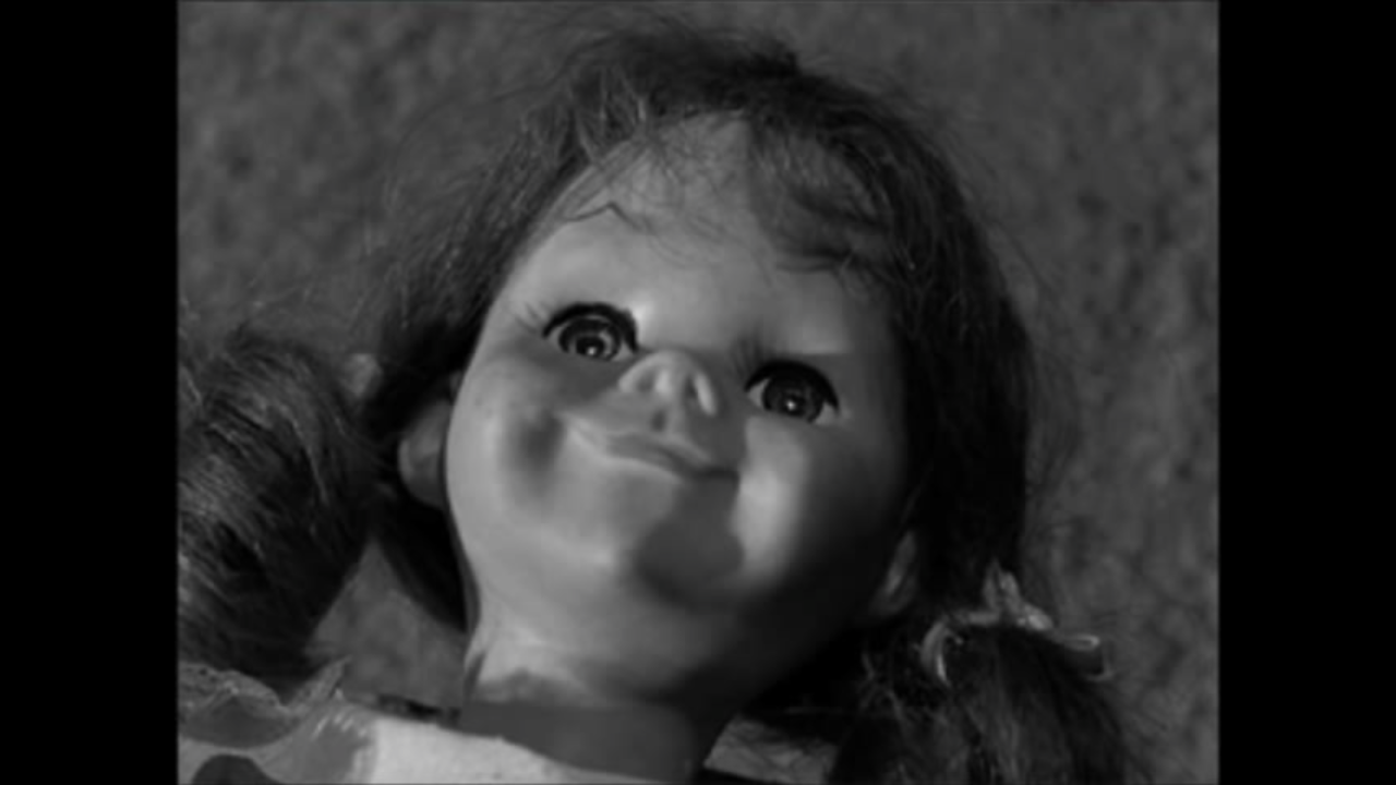 A black and white screen cap from The Twilight Zone. Close up on a doll's face (Talky Tina) looking straight up with an unsettling grin. 