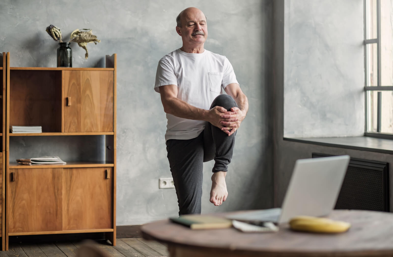 A senior man performing a yoga pose that helps improve hip flexibility and balance in front of his laptop.
