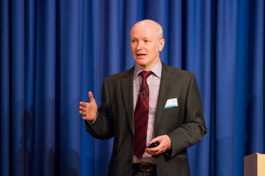 A male-presenting individual in a suit is giving a speech in front of a blue curtain. 