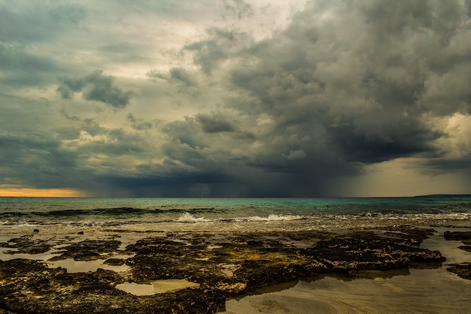 This is an image of dark rain clouds far in the distance over the ocean, as well as a beach covered in seaweed and tidepools. The ocean and weather are both studied through different branches of geoscience.