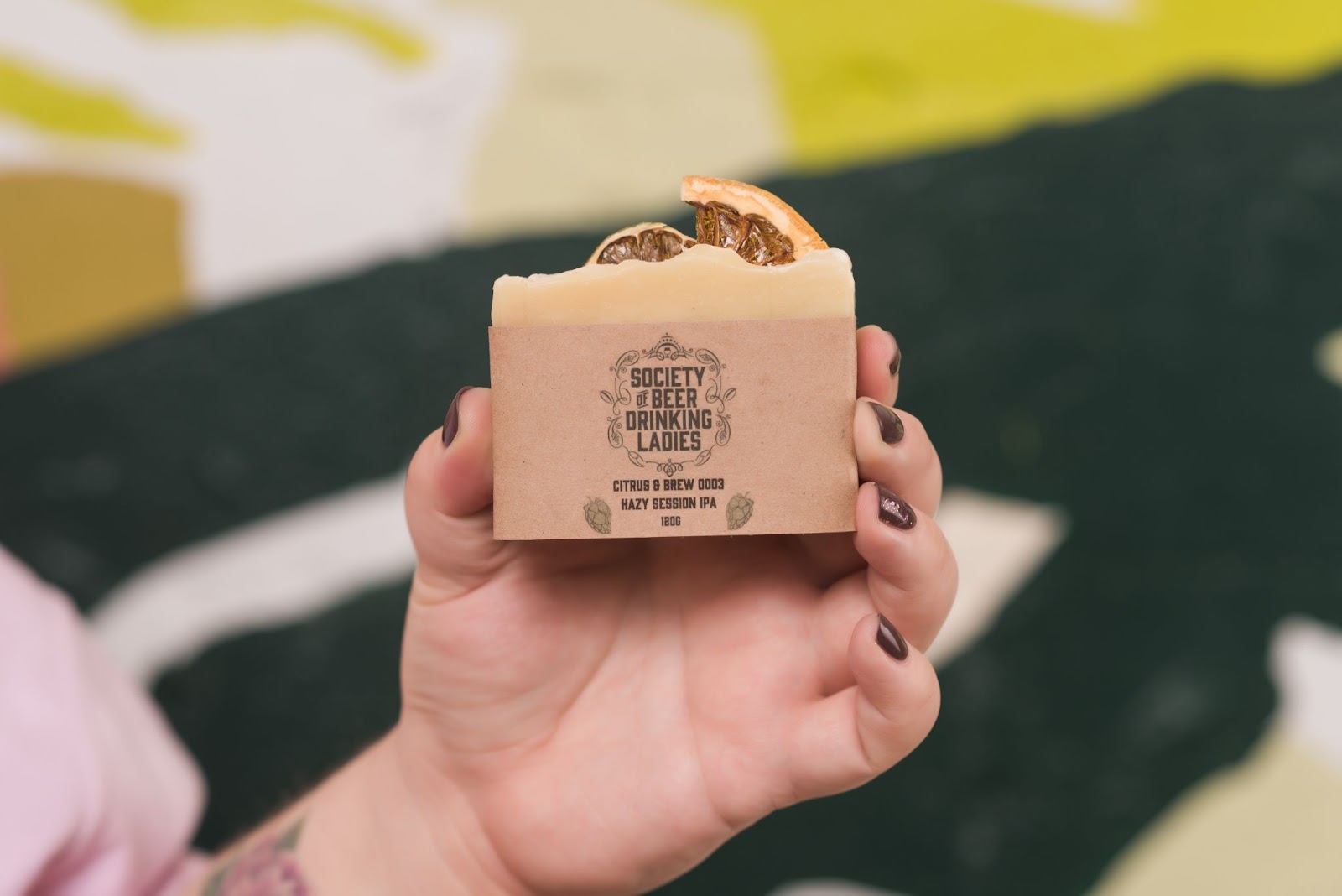 A hand with purple nail polish holding up a light orange bar of soap with the society of beer drinking ladies logo on it. 