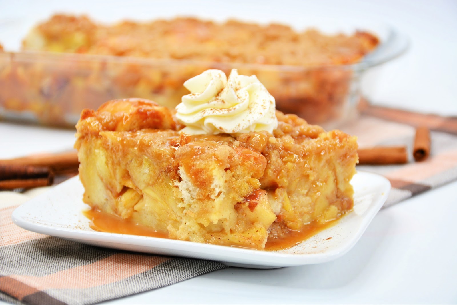 close up of slice of baked apple french toast with whipped cream on top on white plate with cinnamon sticks in background 