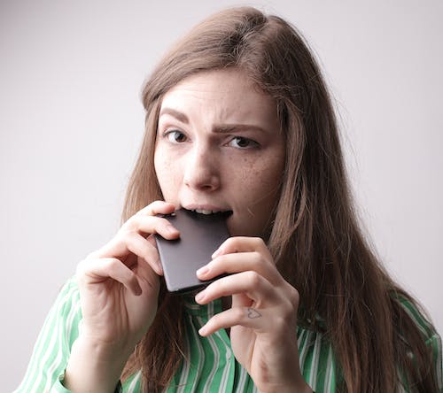 Free Nervous young female in casual clothing standing with cellphone in mouth after unpleasant phone call on gray background in studio Stock Photo