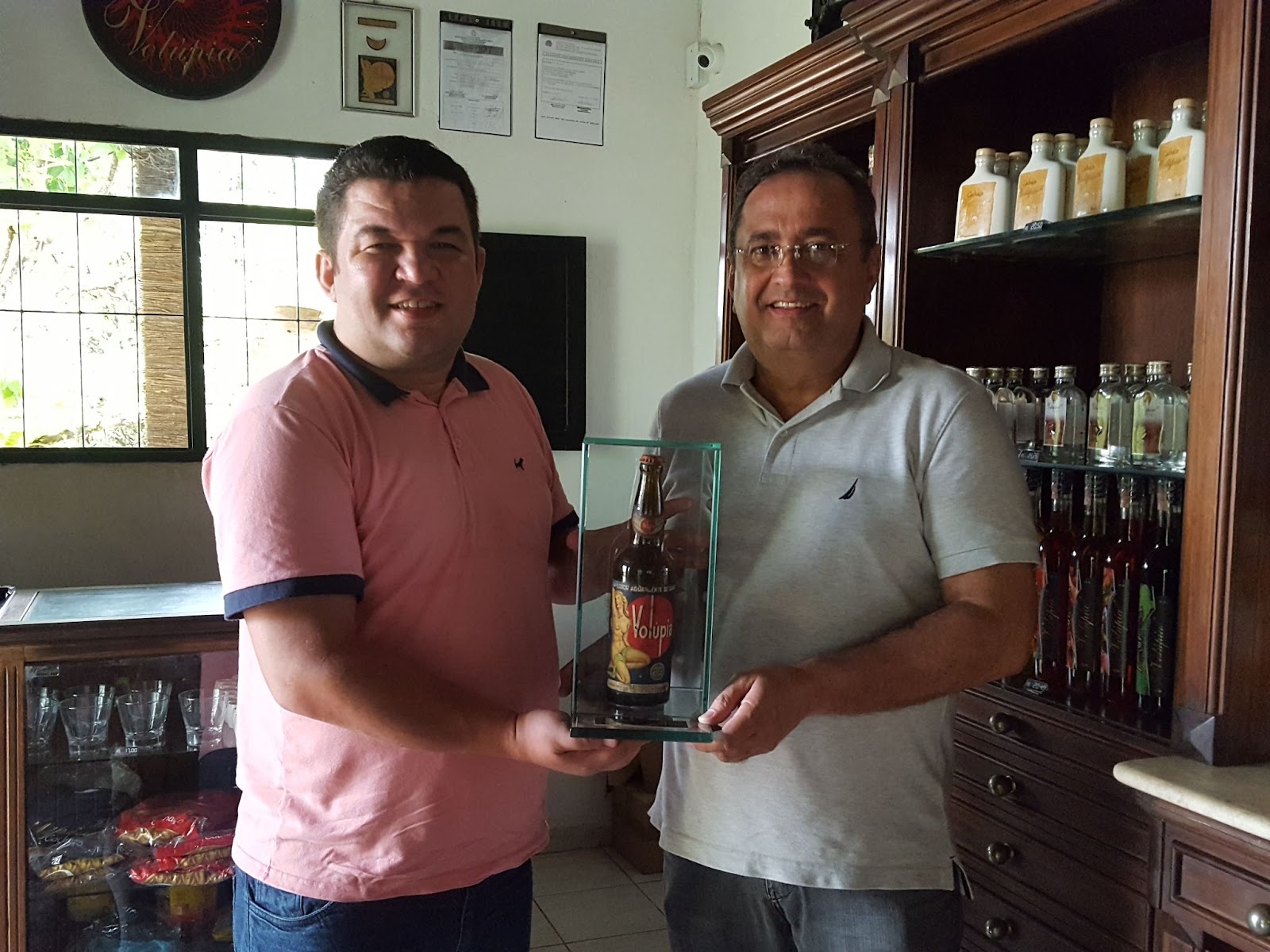 O colecionador de cachaças, Giovani Moser, entrega uma garrafa histórica cor de âmbar para o produtor da cachaça Volúpia. Ao fundo estantes de madeira com garrafas de cachaça. 