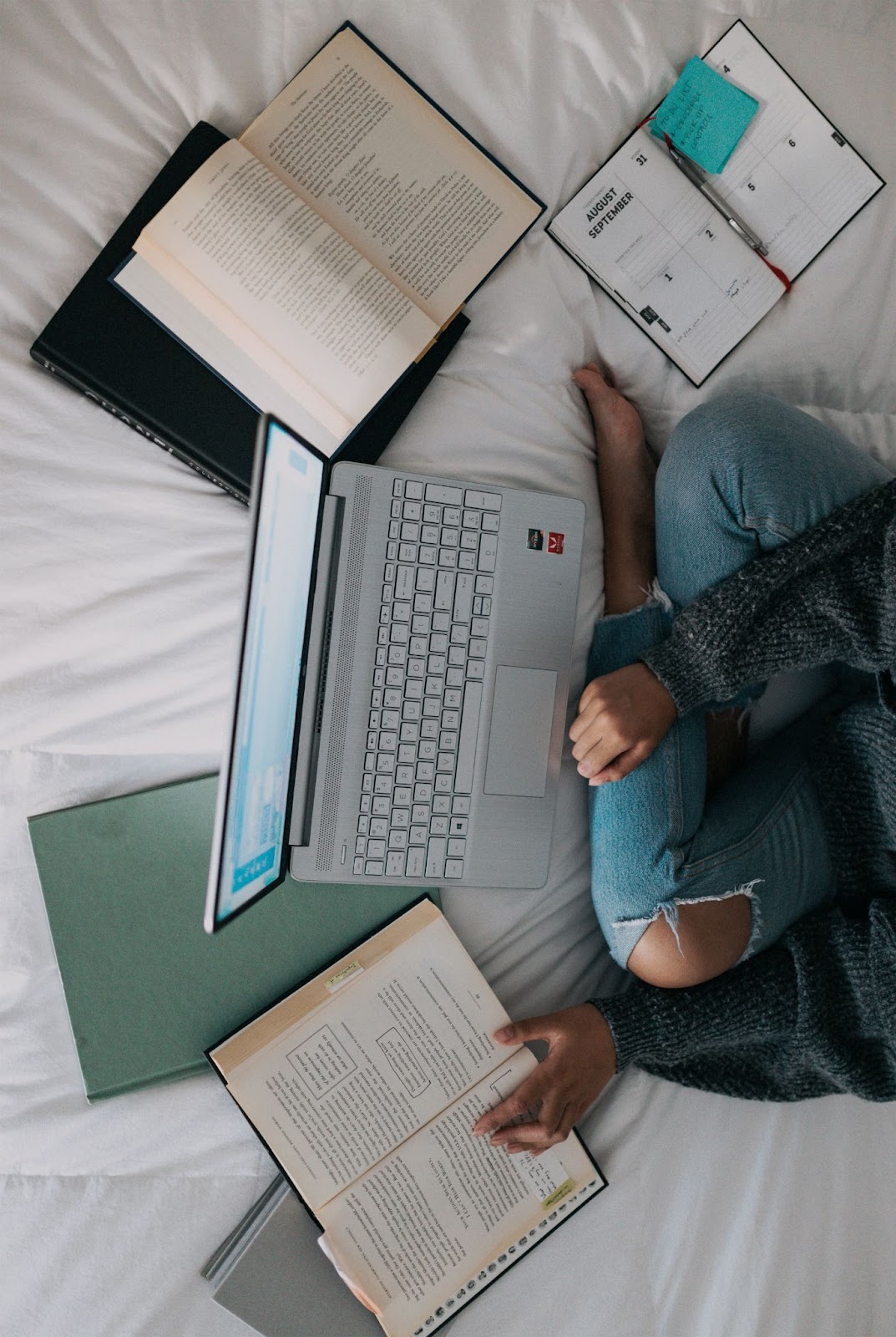An overhead shot of woman sitting cross-legged on a bed with a laptop, several books and a day planner surrounding her.