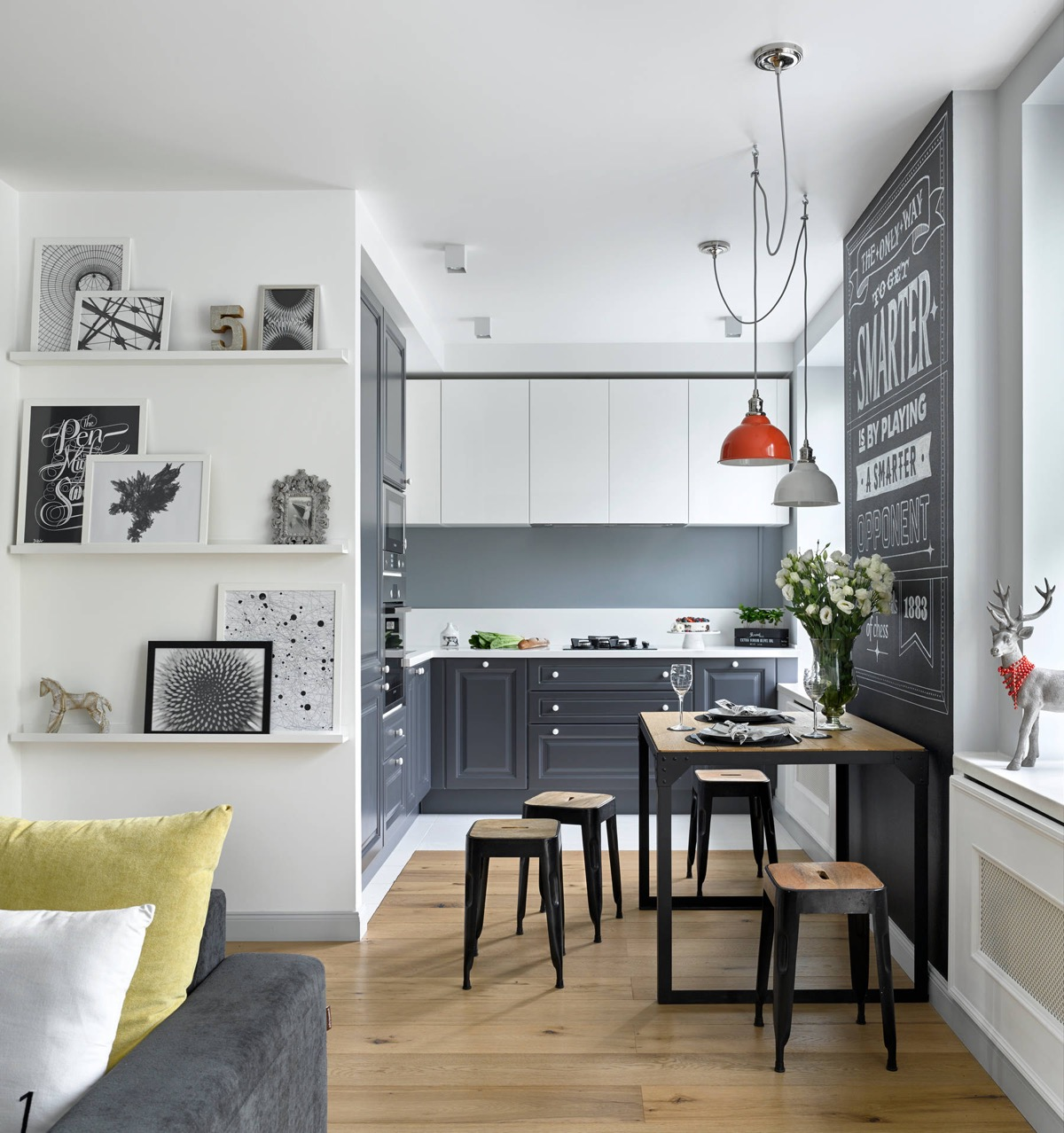 a small kitchen is positioned in the corner of the living room area. grey base cabinets and white upper cabinets give the small space some contrast. a wooden dining table separates the kitchen and living areas