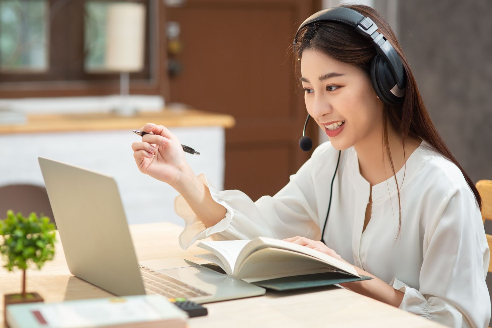 an Asian brown hair woman in a white shirt self-studying on laptop with a headset and an opened book