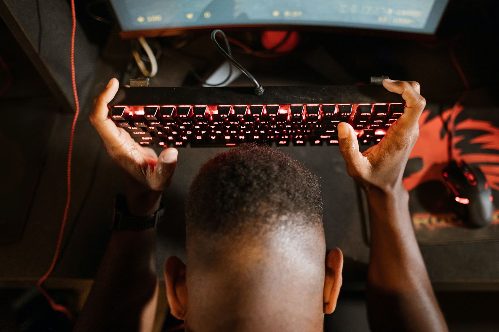 Homem de pele escura segurando teclado de led vermelho em frente a mesa com monitor e mouse.