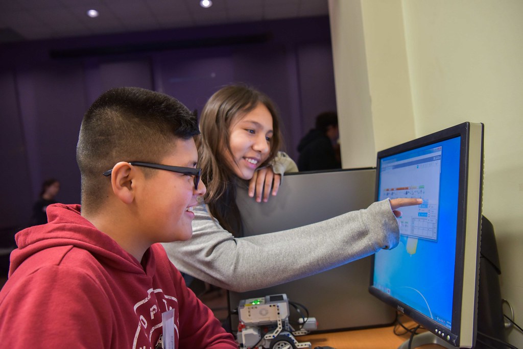 Two high school students share a smile as they discuss something on their computer screen. A young male student with glasses sits at the computer, while a female peer points to something on the screen as she is smiling.