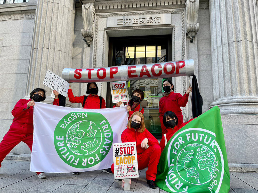 Activists in red boiler suits hold STOP EACOP and Fridays For Future banners outside bank entrance