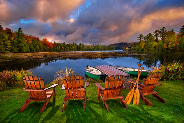 The image shows a peaceful lake surrounded by trees. The sun is setting through clouds. Adirondack chairs and canoe paddles are in the foreground.