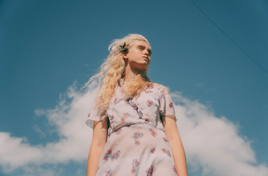 Low angle of young tender female in dress with floral ornament looking away under cloudy sky