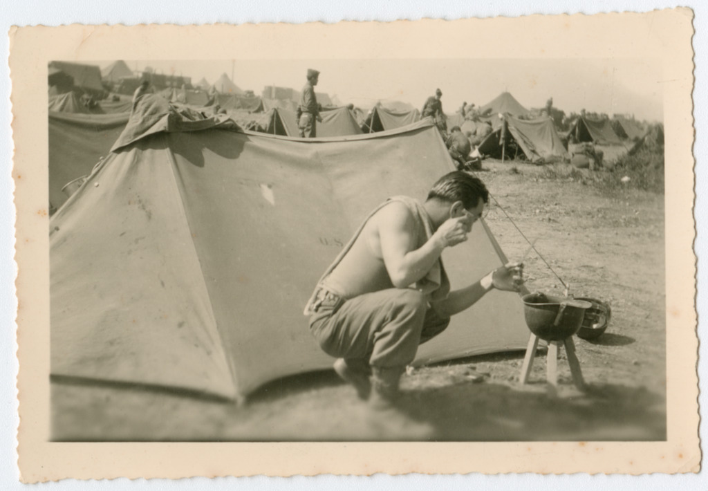 A Nisei soldier crouching to shave next to his tent at a military encampment.