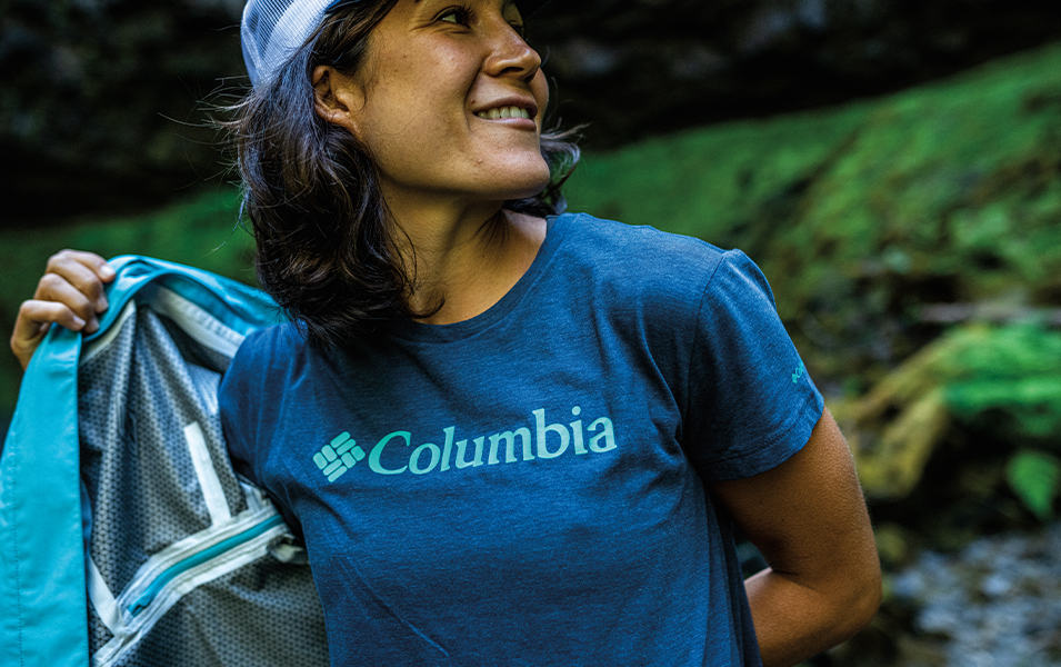 A woman in a blue Columbia Sportswear shirt takes off her rain coat as she smiles and turns her head to the side.