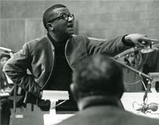 Billy Strayhorn, in a zip-up jacket, talks and points at someone in a crowded room