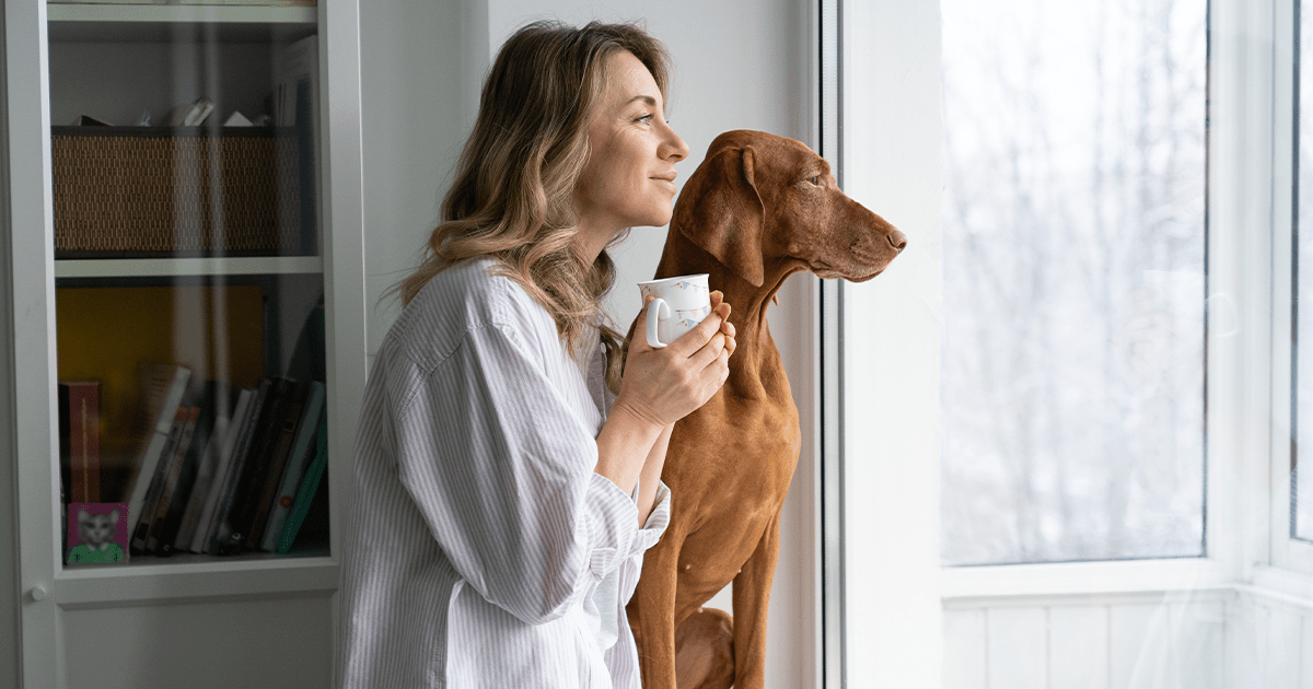 Woman holding cup of coffee standing next to Vizsla dog both looking out a window 