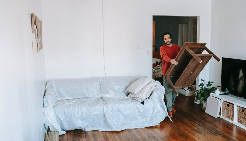 A man is carrying a coffee table into his new apartment. The couch is still wrapped in a protective covering from the move.