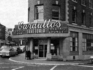 Black and white photo at street level of a three story brick building. Cars on the left. Sign above glass doors reads "Cardullo's" in large letters
