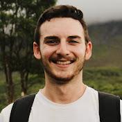 A young man with a light beard, brown hair, white tshirt and rucksack smiles at the camera. He is outside, there is a tree in the background, some fog and a green field.