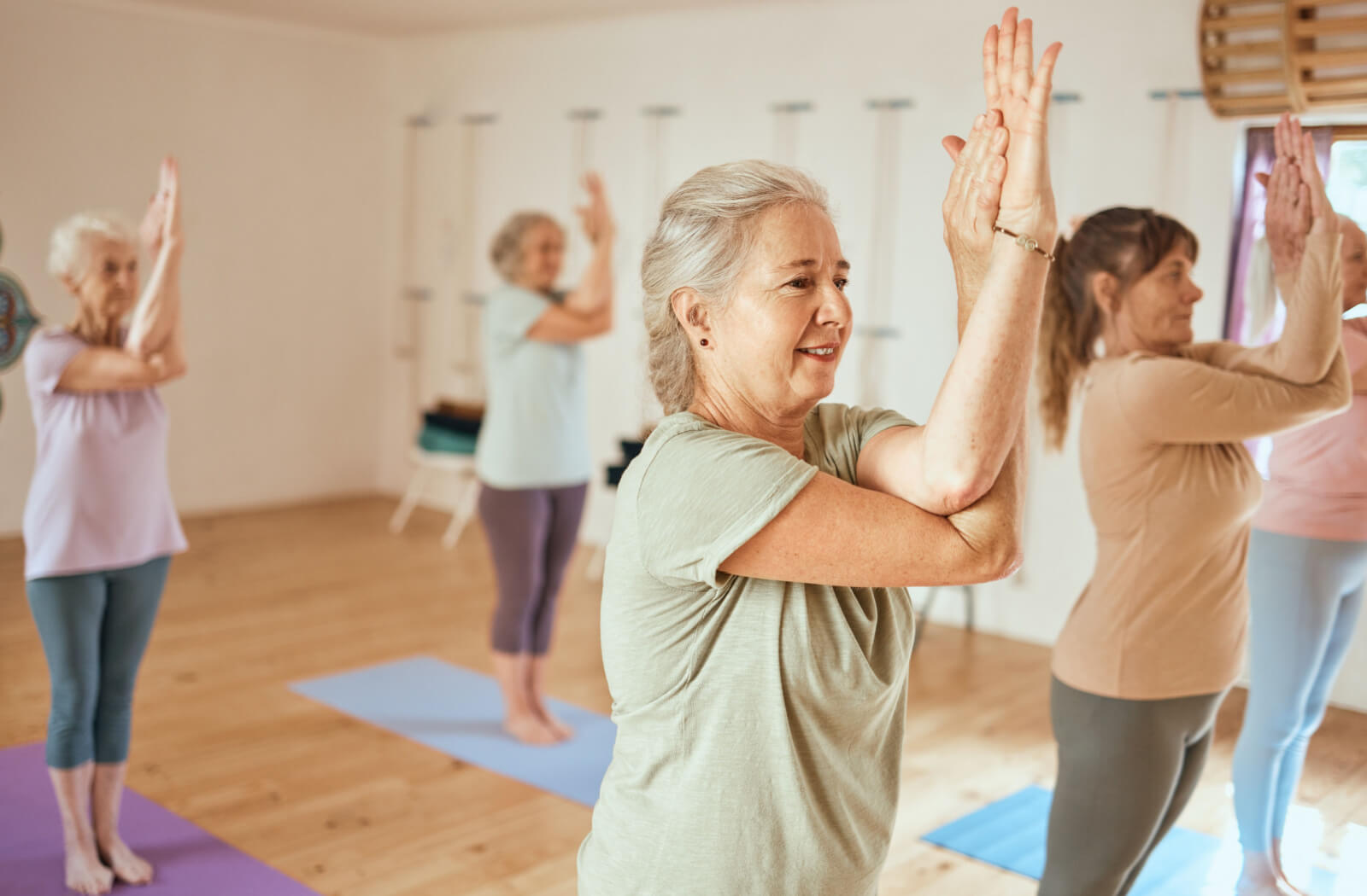 A group of women standing on a yoga mat doing balance exercise in a  memory care facility.