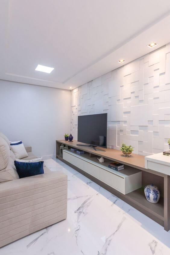 Sala com piso porcelanato reproduzindo mármore branco, revestimento 3D branco na parede de fundo, rack comprida amadeirada com TV e sofá branco com almofadas azuis.