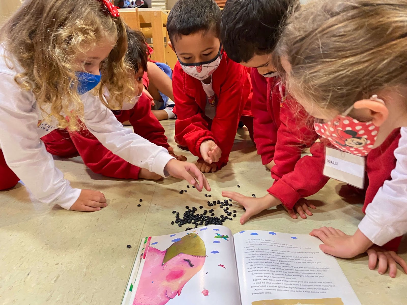 A imagem mostra crianças em torno de um livro e feijões.