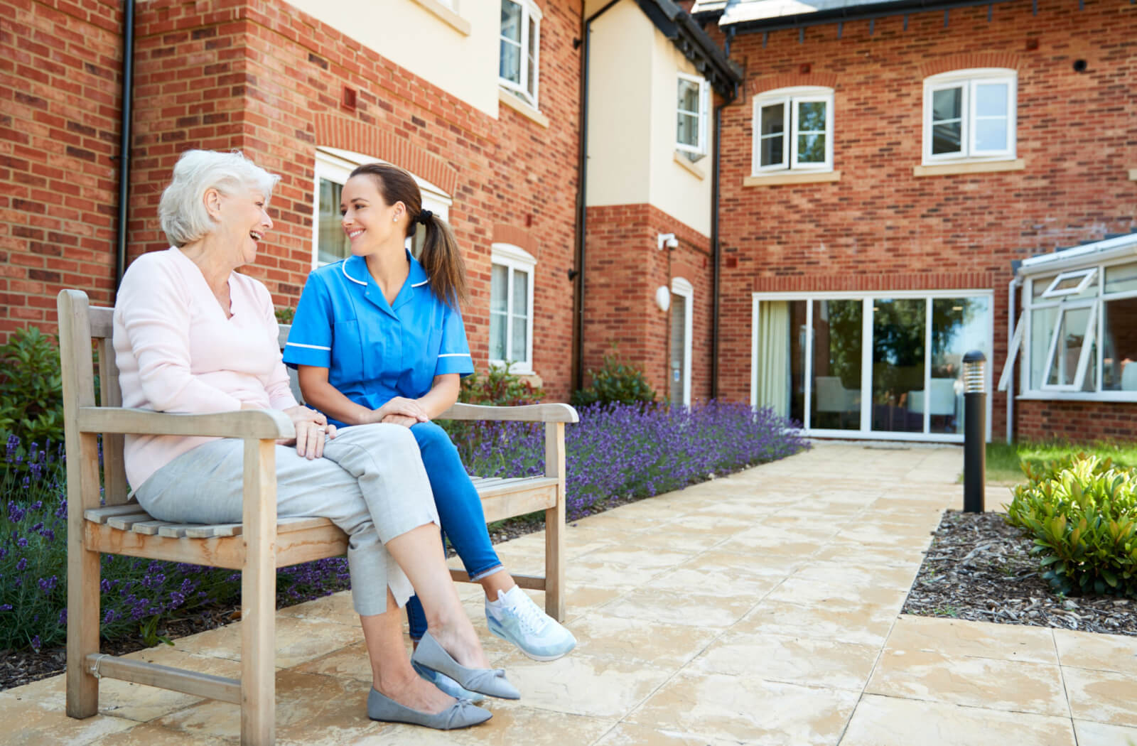 A senior woman with a friendly expression on her face in a senior living facility sitting on a bench talking to her nurse.