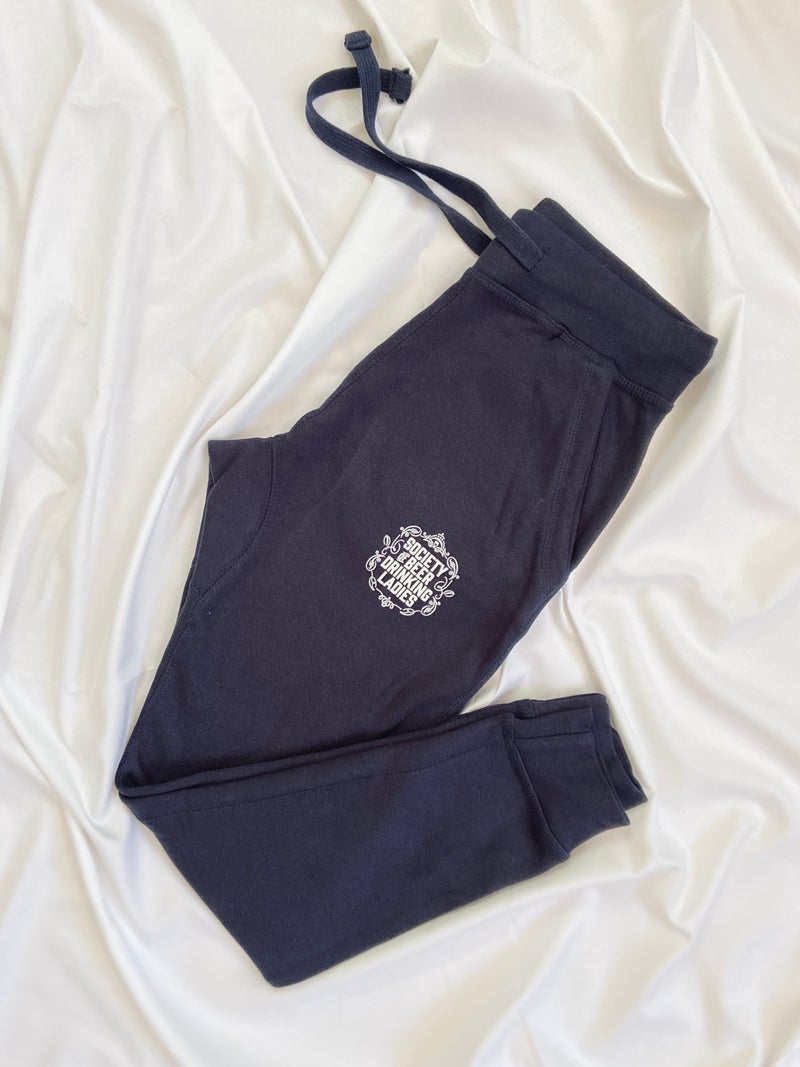 Navy blue sweatpants with a white Society of Beer Drinking Ladies Logo on the leg.  They are lying folded at an angle on a white sheet. 