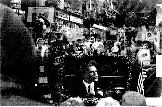 Black and white newspaper photo of a TV screen with a person speaking surrounded by a small  United States flag and small containers.