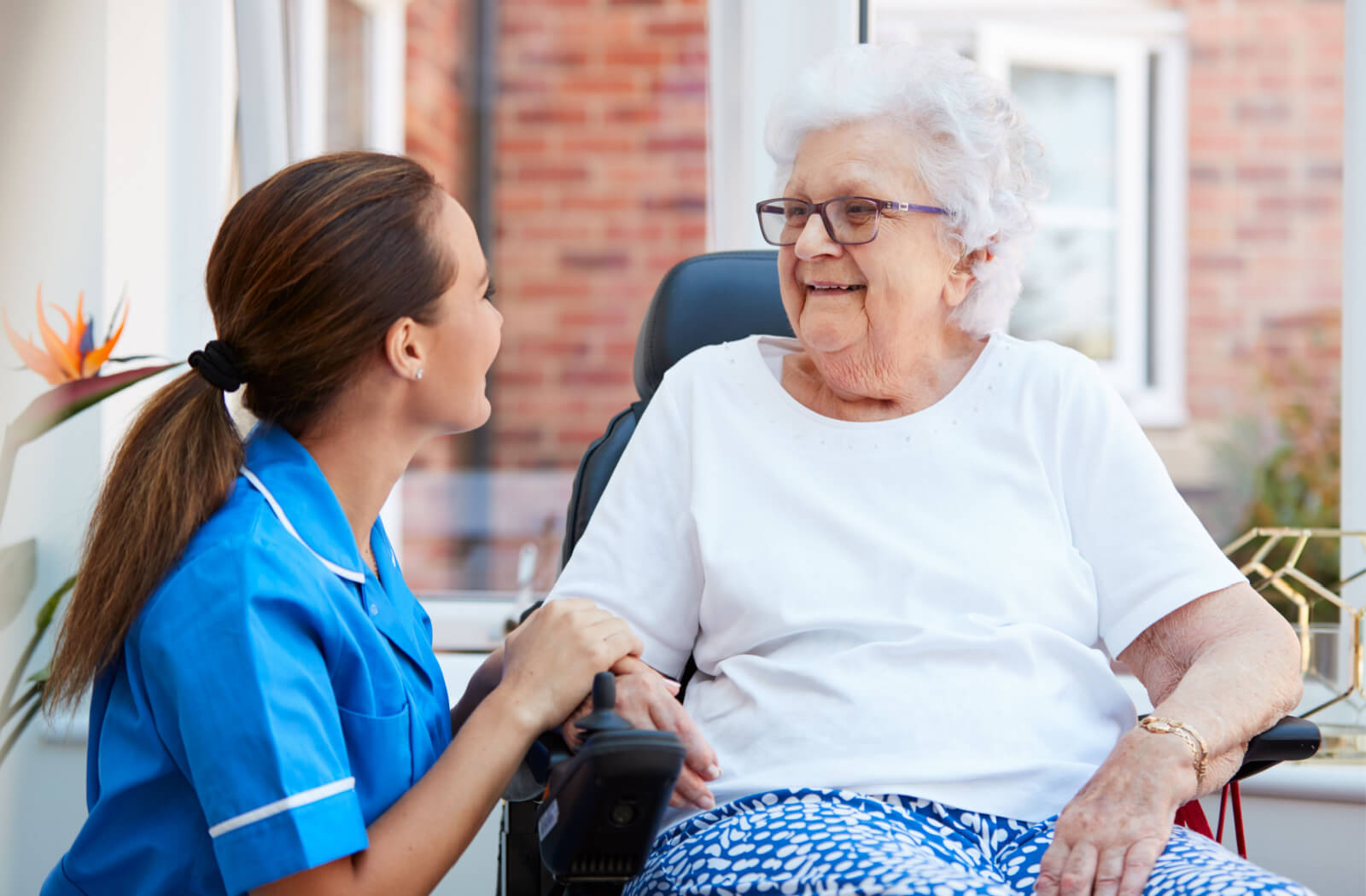 A senior woman in a senior living facility sitting on a chair smiling and having a conversation with a nurse.