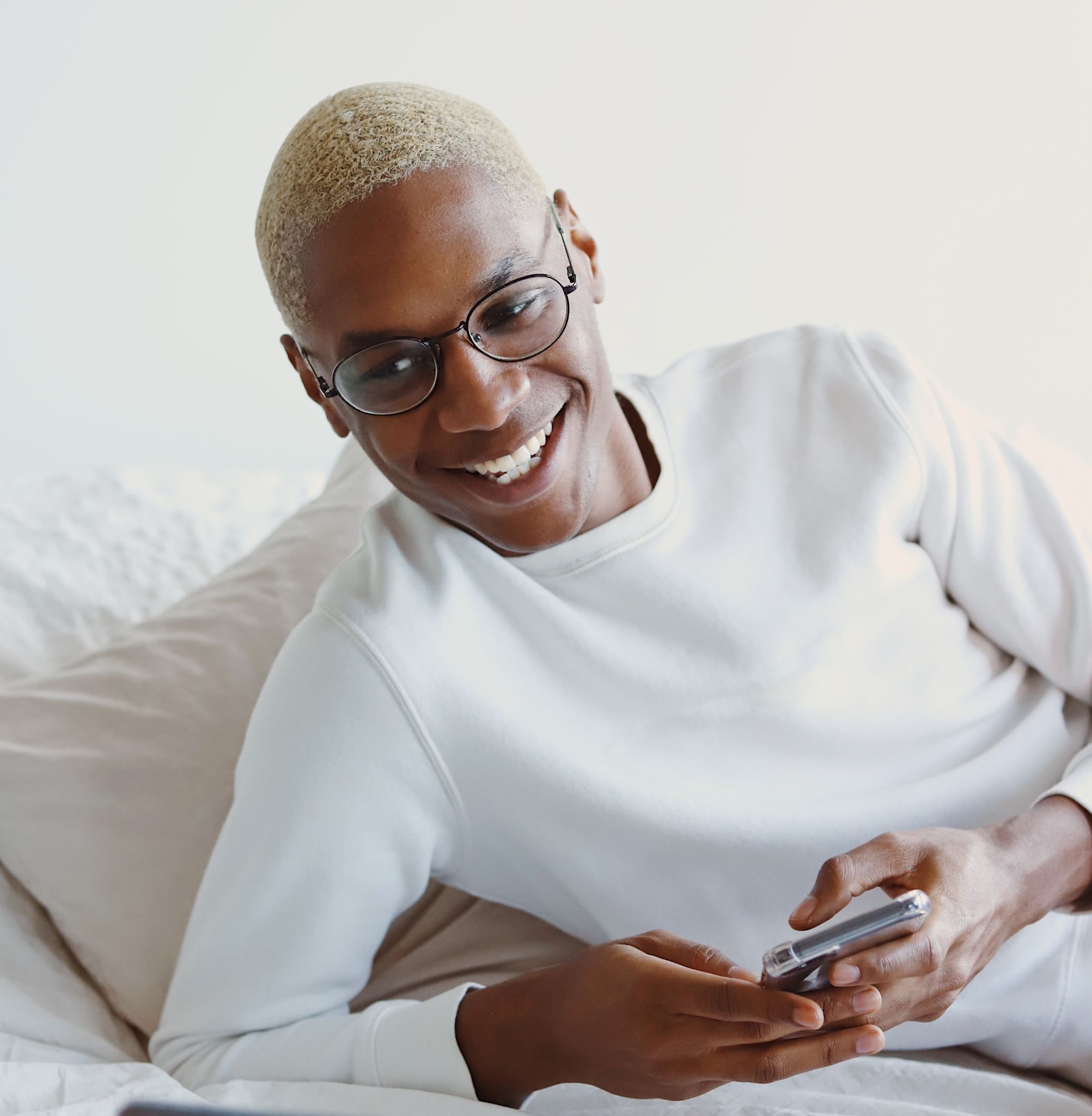 A picture of a young man, smiling while looking at his phone while lying on a bed