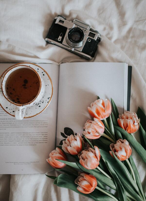 foto de uma xícara de chá, câmera fotográfica, um livro aberto e um buquê de flores organizados em uma cama